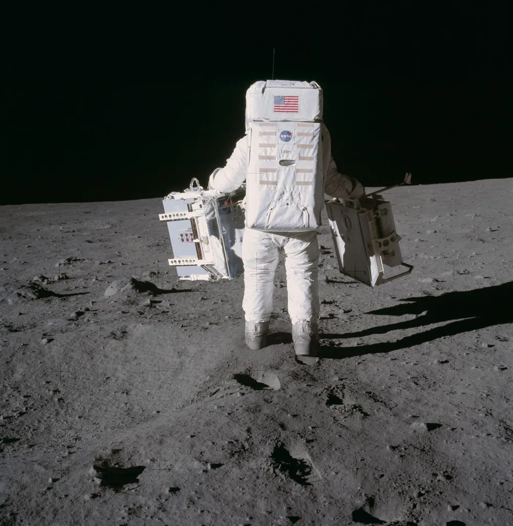 Astronaut Buzz Aldrin seen from behind on the gray lunar surface carrying two science packages, one in each hand