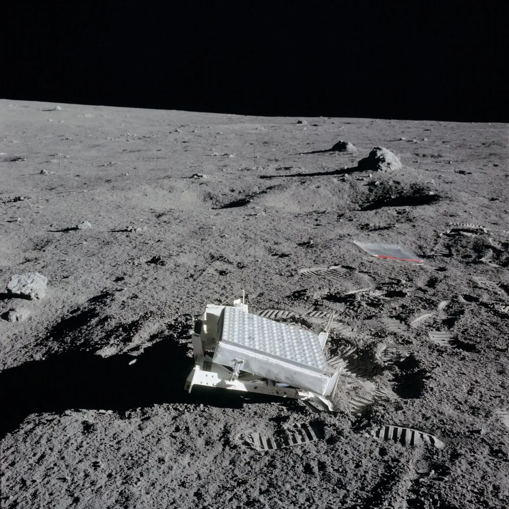 A rectangular metal retroreflector array covered in round prisms, sitting tilted on gray lunar soil among footprints