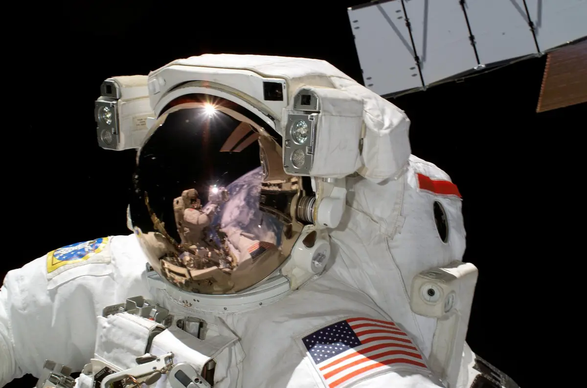 Astronaut in a white spacesuit during a spacewalk, with Earth and a second astronaut reflected in the gold visor and a solar array behind
