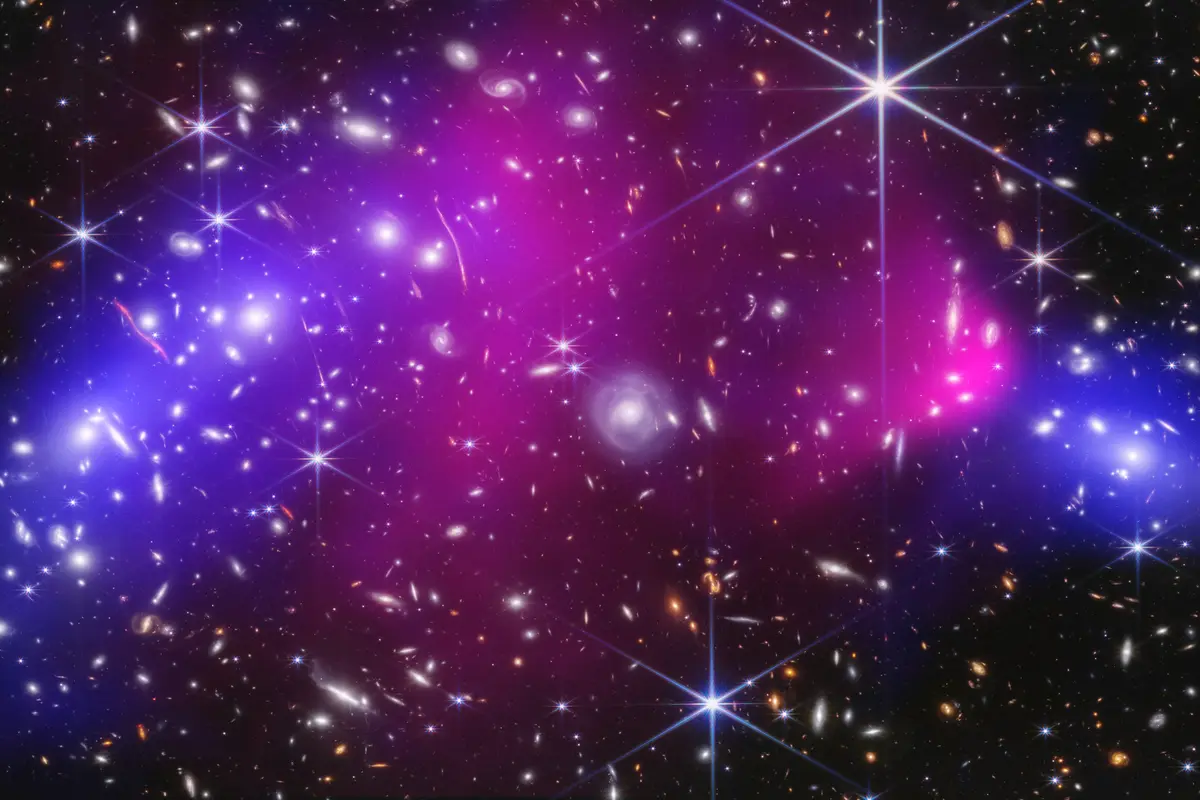 Two colliding galaxy clusters seen by Webb and Chandra: hot gas glowing pink in the middle and mapped dark matter glowing blue on both sides, among thousands of galaxies and stars