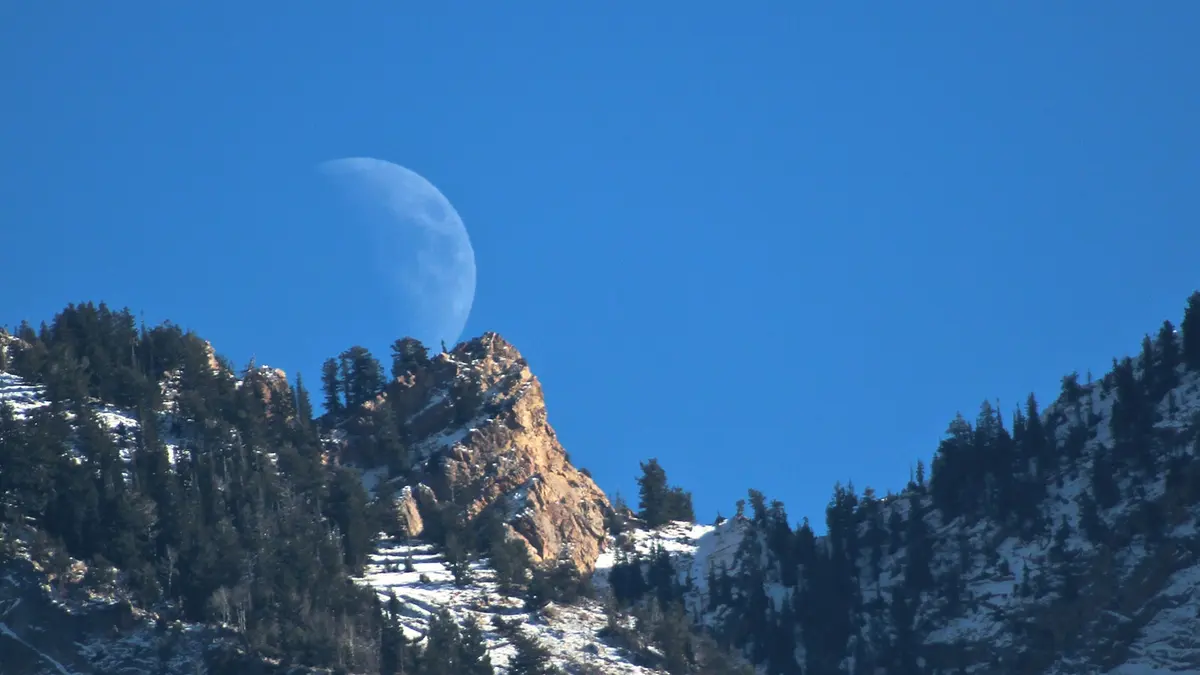 A pale gibbous Moon in a deep blue daytime sky rising behind a rocky, snow-dusted mountain ridge in Utah