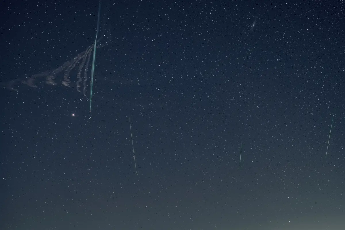 A bright green Leonid meteor trail in a dark blue sky, with a twisting smoke train beside it and a few fainter meteors