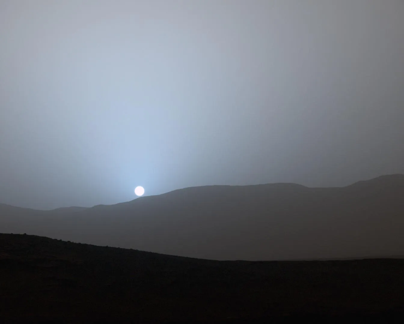 The Sun setting behind low hills on Mars, surrounded by a soft blue glow in a gray sky