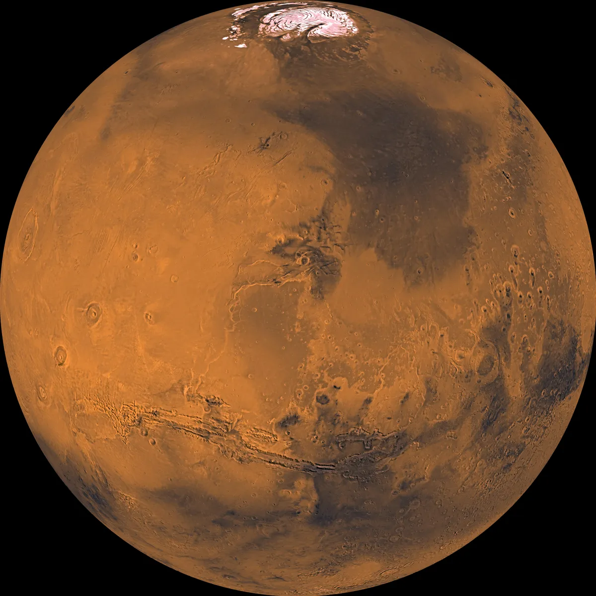 Mars seen as a full globe, orange-red with darker patches, a long canyon across its middle and a white polar cap at the top