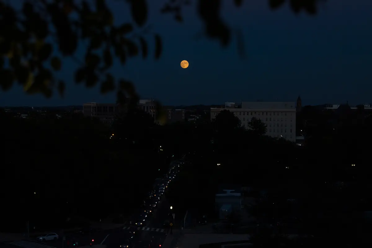 The nearly full Moon low over the rooftops and trees of Washington at dusk, with traffic below