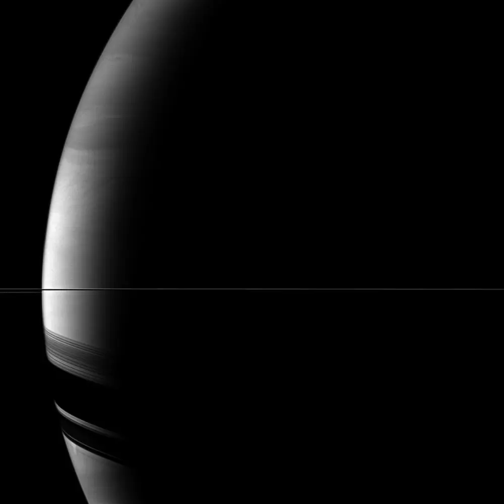 Saturn's rings as a bare thin line across the frame, with broad dark ring shadows on the planet's southern hemisphere