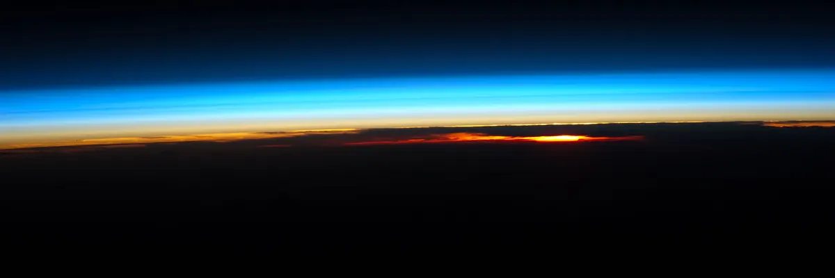 The red-orange Sun setting at the edge of Earth, under a bright blue band of atmosphere that fades into black space