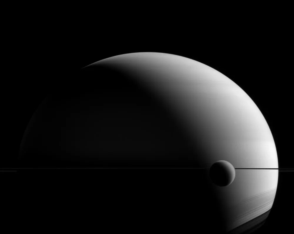 Saturn's huge hazy crescent with its moon Titan, a small round disk, passing in front near the thin line of the rings